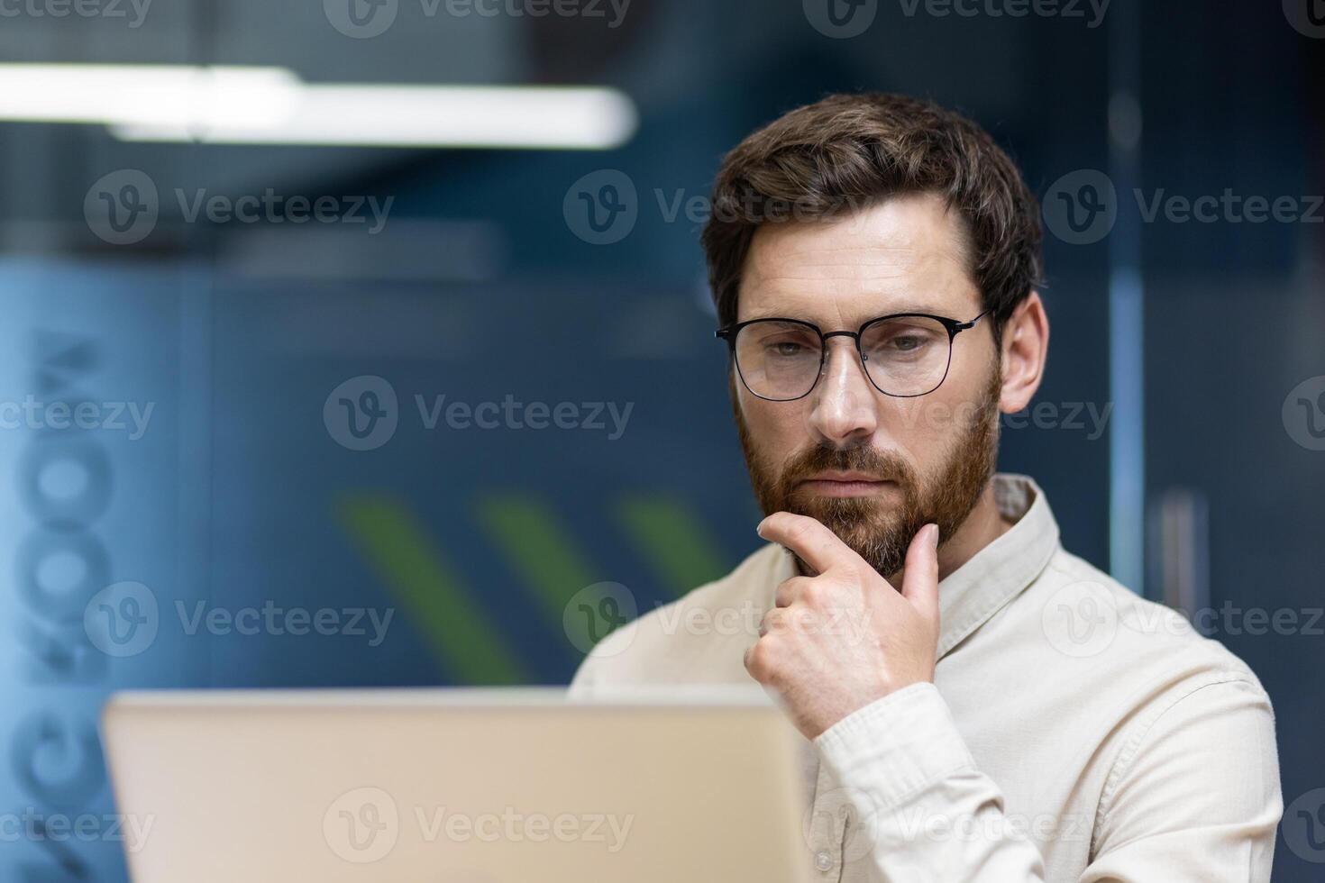 Close-up photo of a thoughtful young businessman sitting in the office at the workplace and looking at the laptop screen while rubbing his beard with his hand.