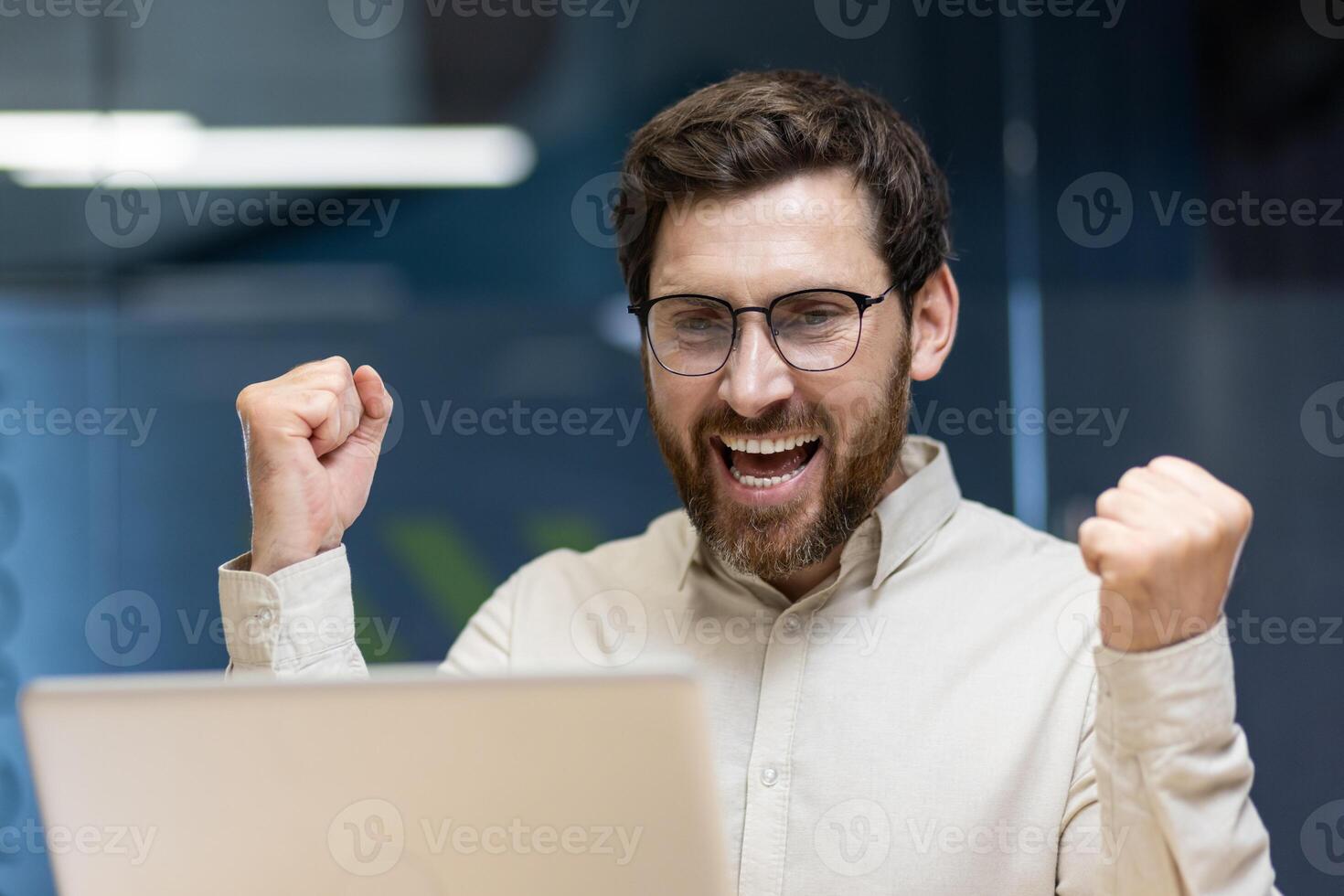 Close-up photo of a young happy man who is in the office, looking at the laptop screen and is happy, showing a victory gesture with his hands.