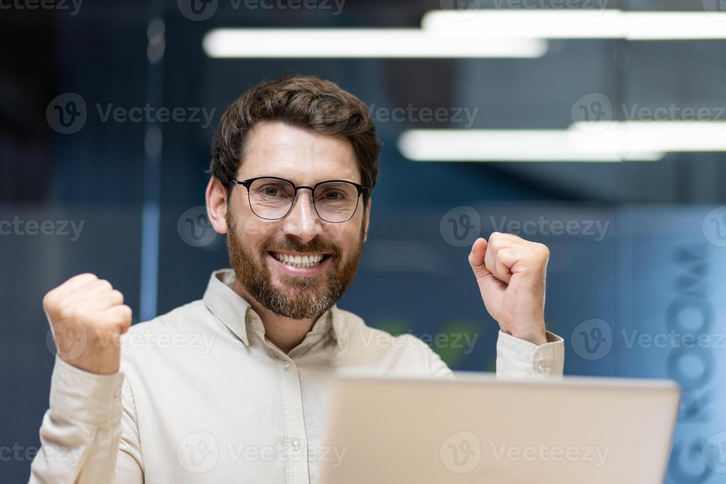 Close-up portrait of a young happy businessman who is in the office, sits at the workplace with a laptop, rejoices at the success, shows a victory gesture with his hands. photo