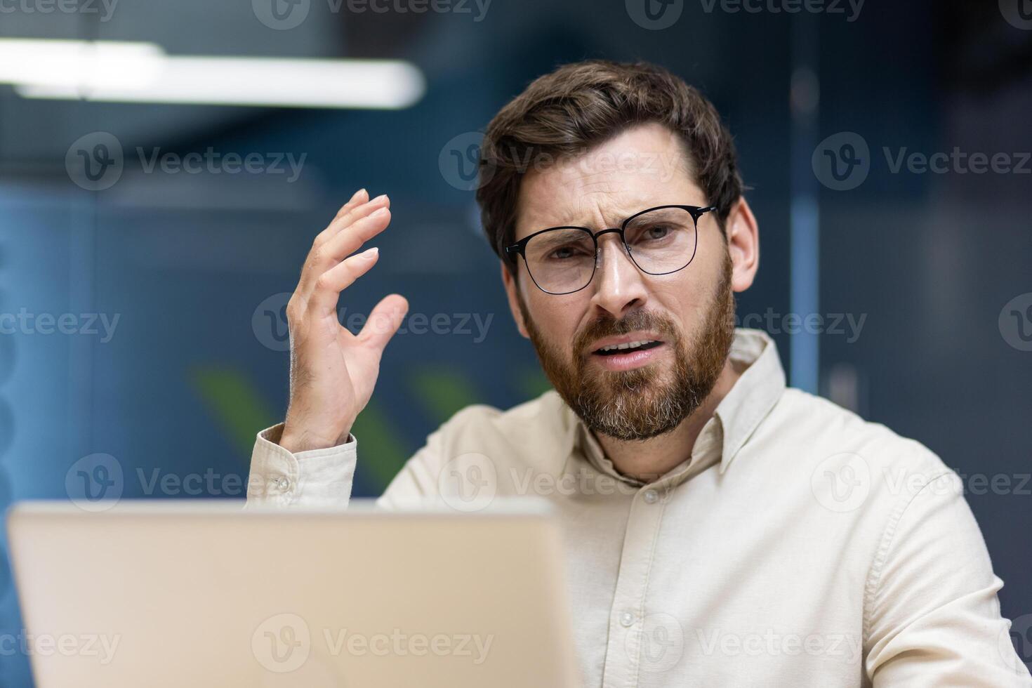 Close-up portrait of an angry and frustrated young male office worker sitting at a workplace with a laptop and looking annoyed at the camera while gesturing with his hand. photo