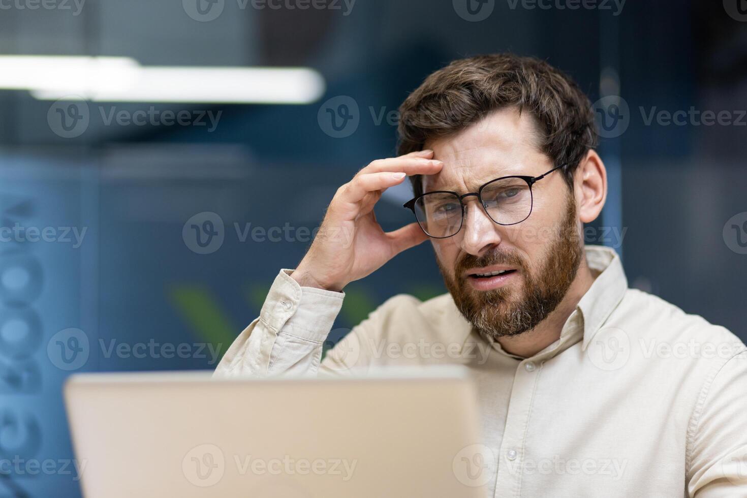 Close-up portrait of a young upset male businessman sitting in the office in front of a laptop and holding his head in frustration. photo