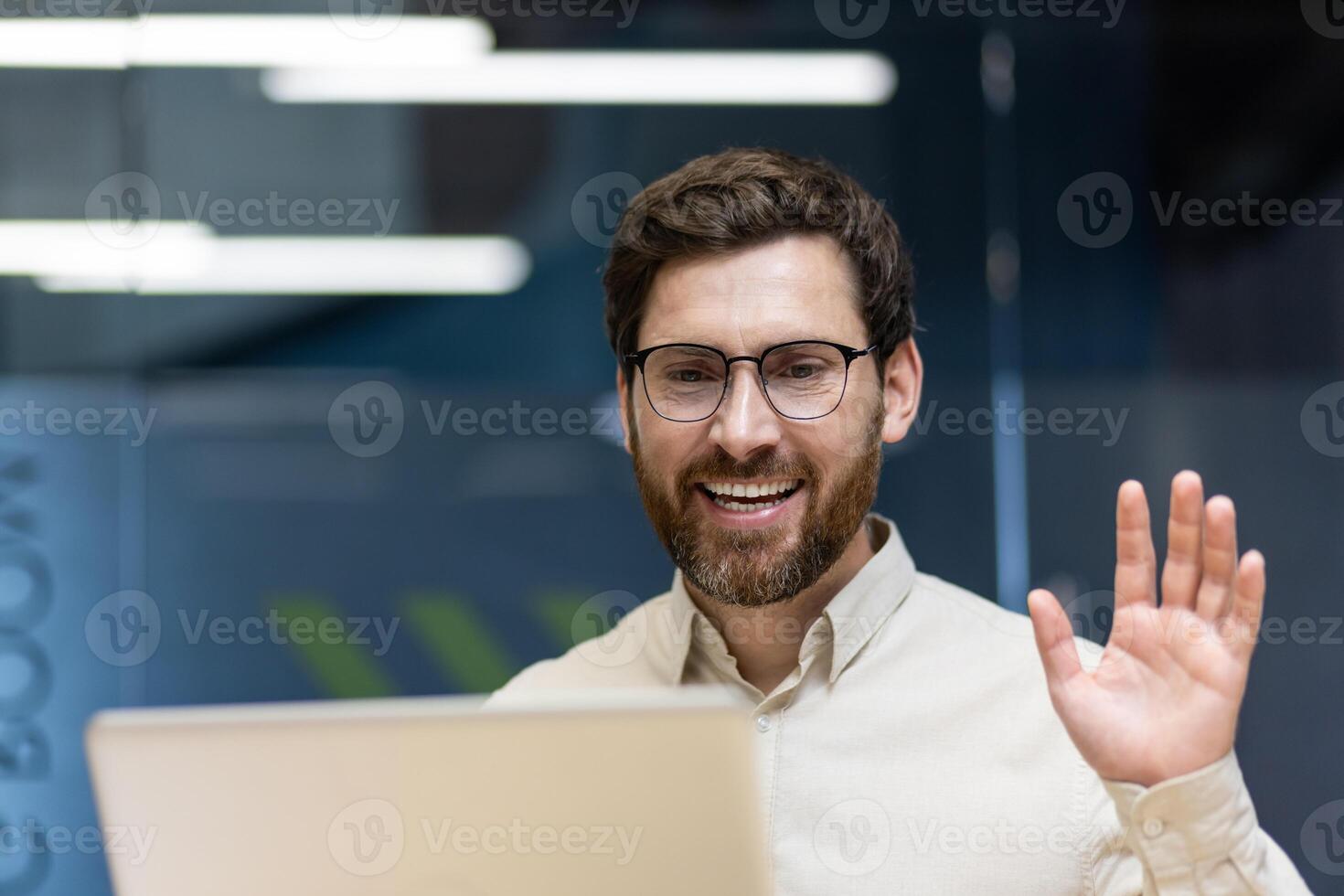 Close-up photo of a young smiling man sitting in an office at a workplace in front of a laptop, greeting and waving at the camera.