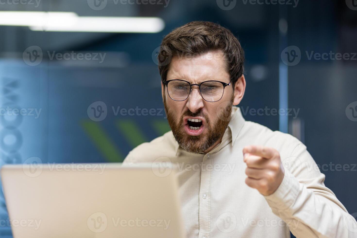 Close-up photo of a young angry man sitting in the office at the desk and shouting while pointing his finger at the laptop screen.