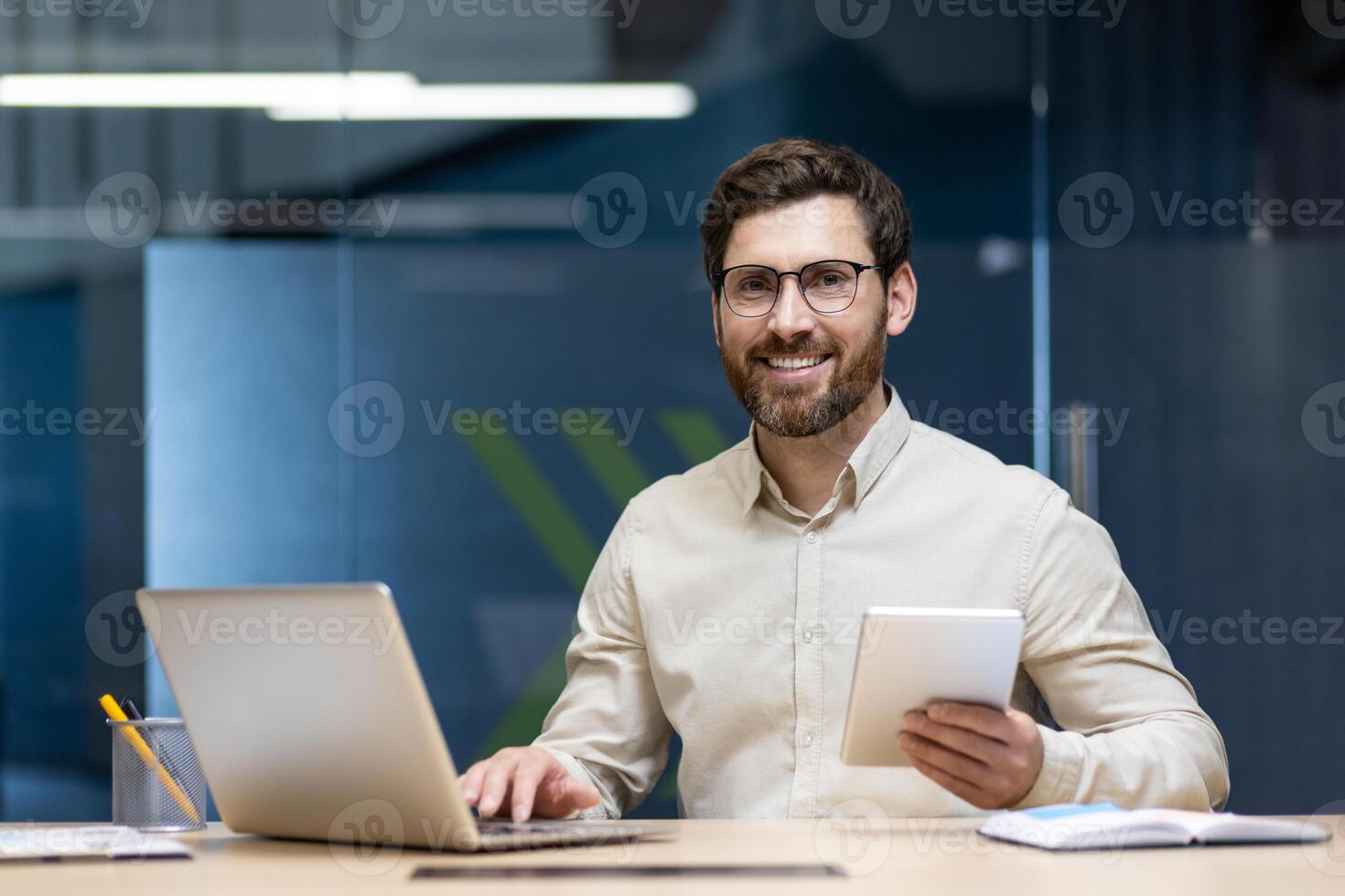 Portrait of a smiling young man in glasses sitting in the office at a desk with a notebook, holding a notebook and looking at the camera. photo