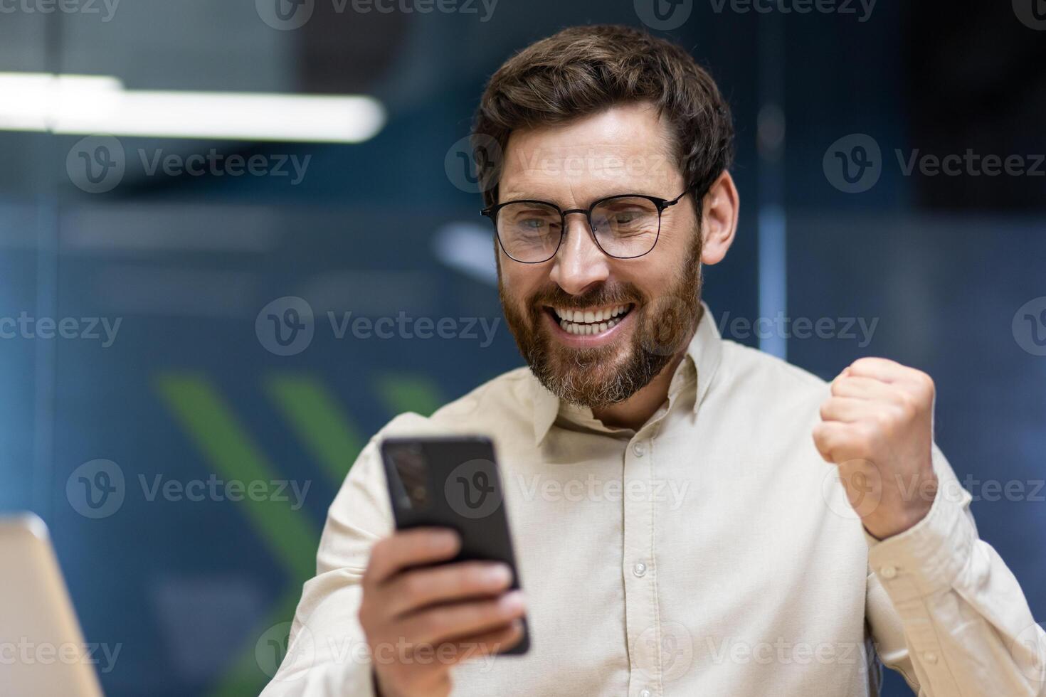 Close-up photo of a young happy man sitting in the office at the table and rejoicing in success, looking at the mobile phone screen and showing a victory gesture with his hand.