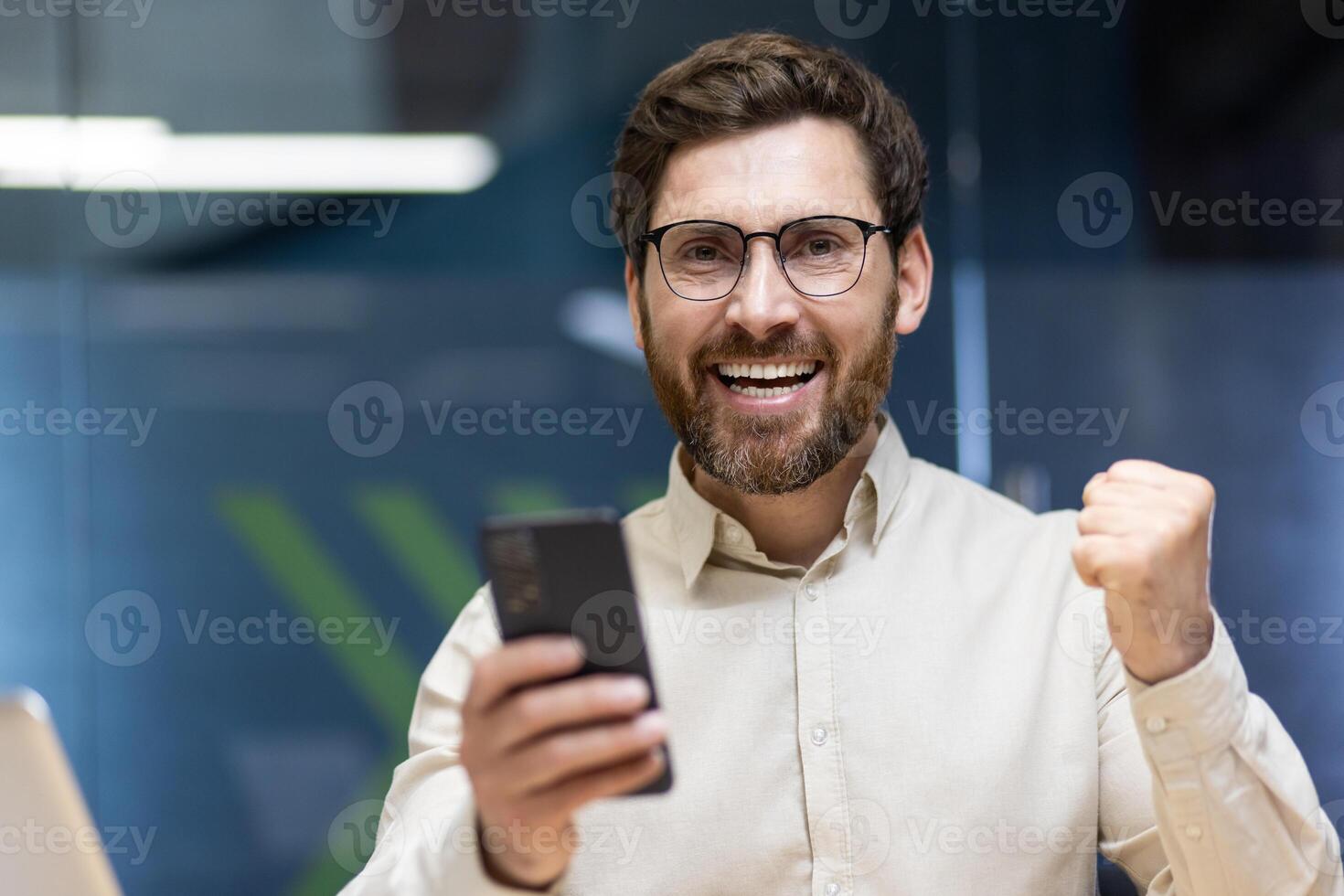 Close-up portrait of a young happy man in glasses and a light shirt who is in the office, rejoices in success and victory, holds a phone in his hand. photo