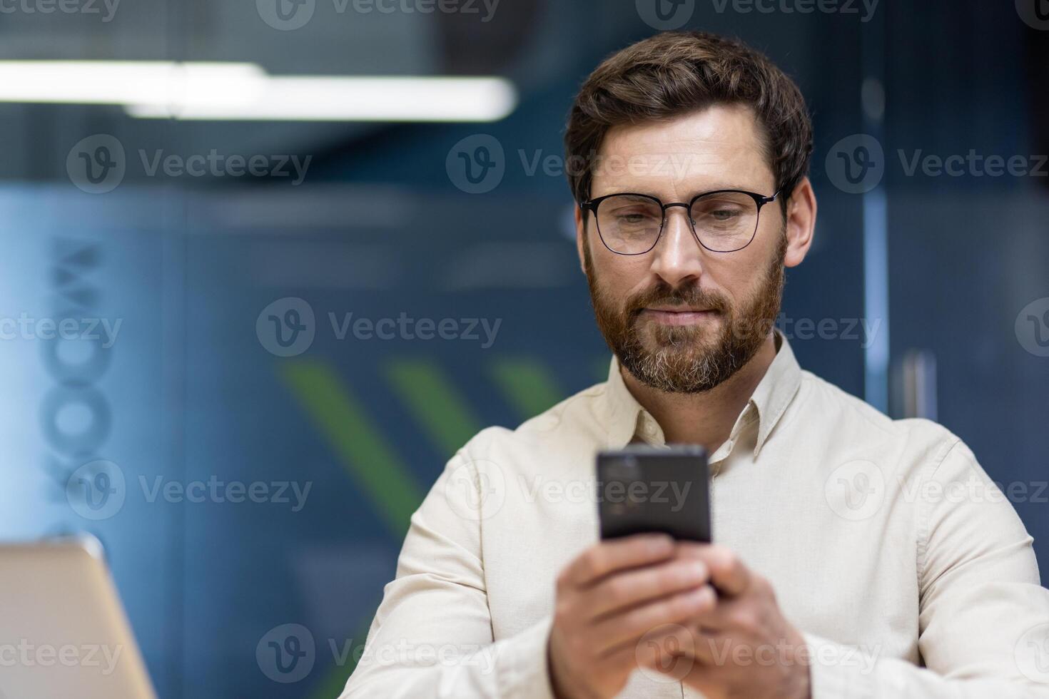 Close-up photo of a smiling young man in a shirt and glasses who is in the office and ,using a mobile phone in his hands.