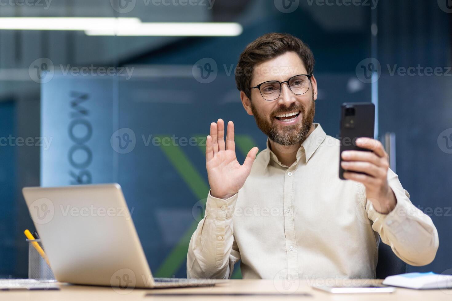A young businessman man is working in the office on a laptop, sitting at a desk and talking on a call through a phone camera, greeting and waving. photo