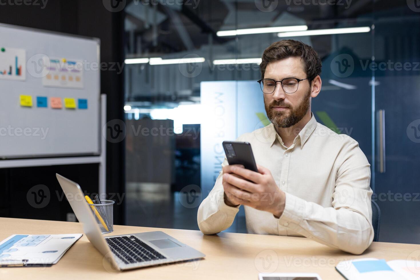 Serious and focused young man sitting in office at desk with laptop on glass sticker board background, holding and using mobile phone. photo