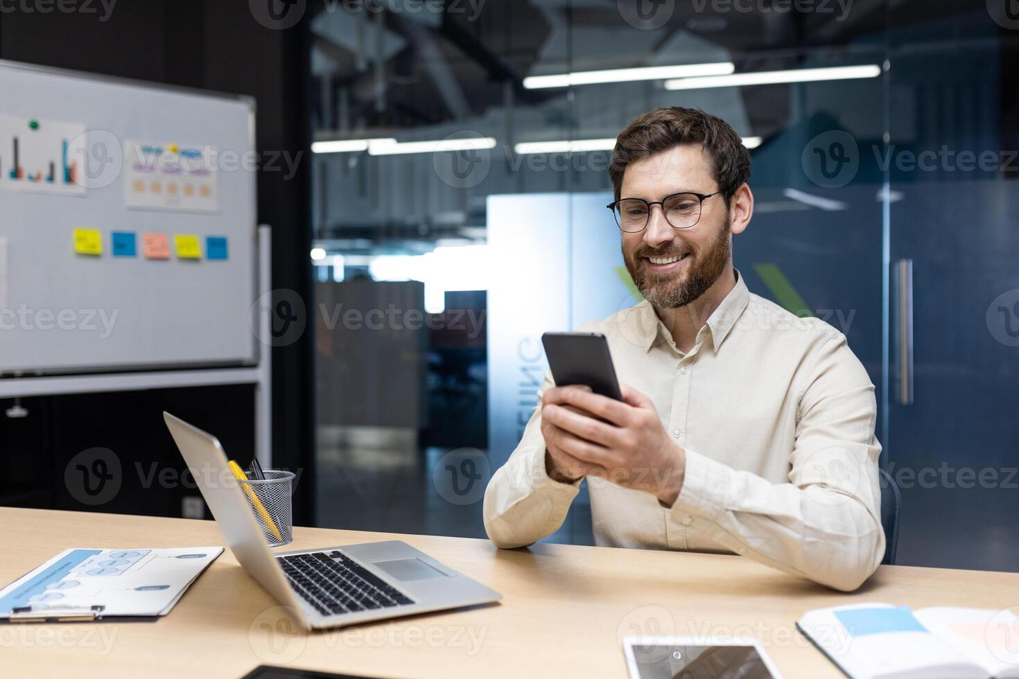 A smiling young man works in the office on a notebook, sits behind a desk on a whiteboard with stickers and uses a mobile phone in his hands. photo