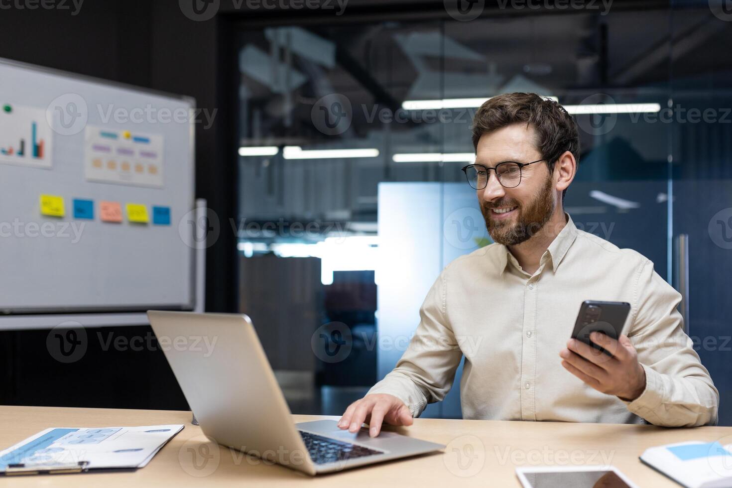 Smiling young man businessman, developer works in office at laptop on whiteboard with stickers, types on keyboard, holds mobile phone in hand. photo