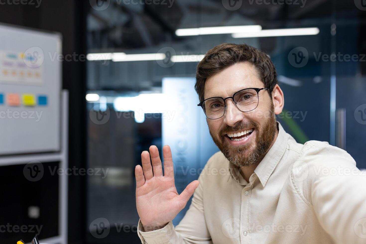 Close-up photo of a smiling young man in glasses and a shirt who is in the office, talking to the camera he is holding in his hand on a call, greeting and waving.