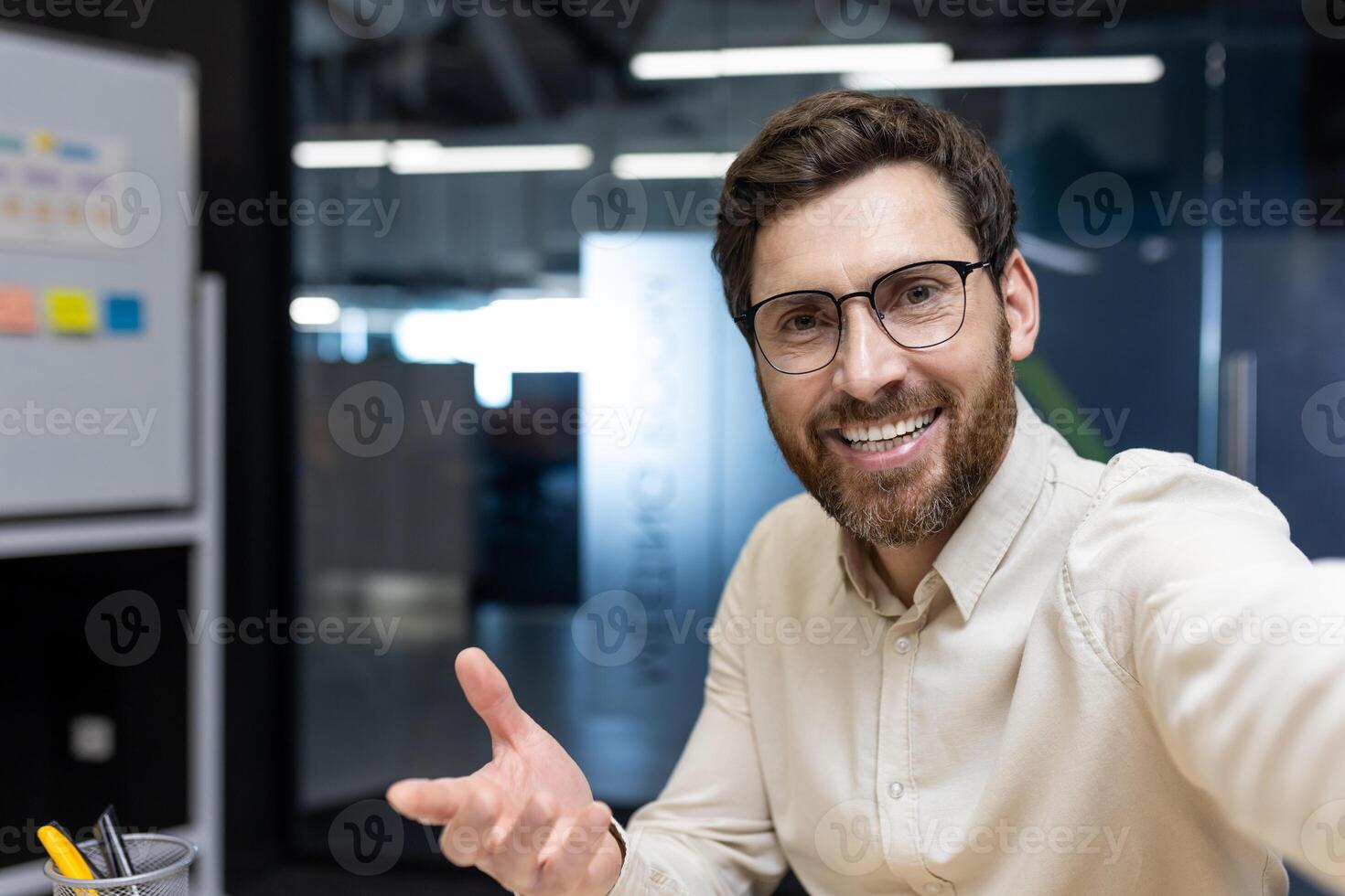 Close-up photo of a young smiling man in a shirt and glasses who is in the office, holding a camera in his hands and talking on the Internet, gesturing to the camera with his hand.