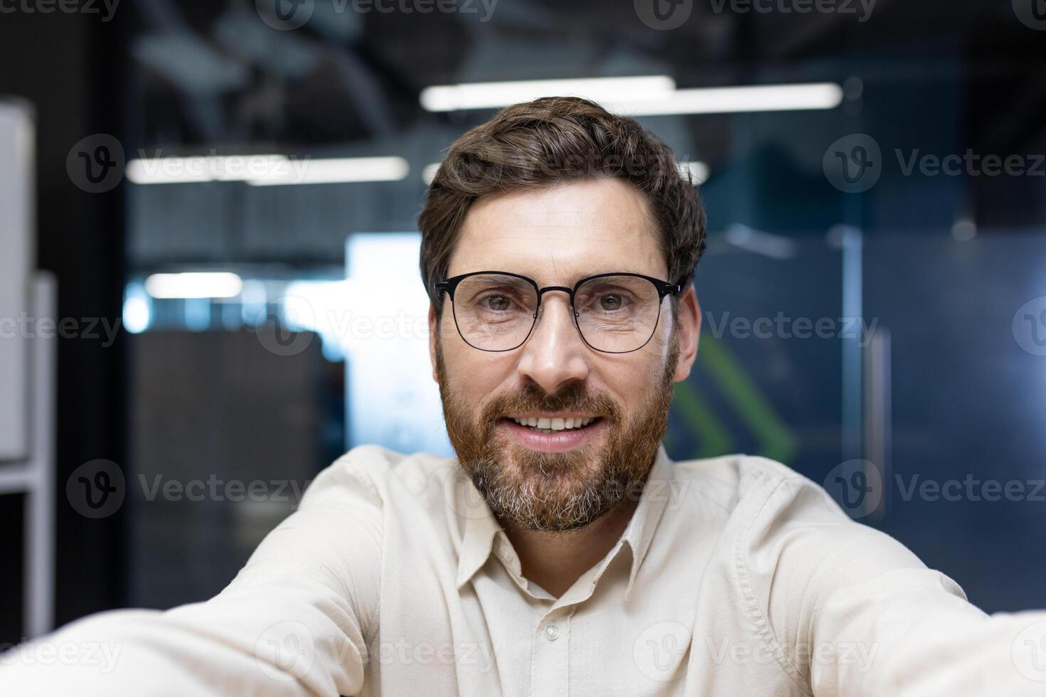 Close-up portrait of a young smiling man with glasses and a beard sitting in the office, talking and looking at the camera he is holding in his hands. photo