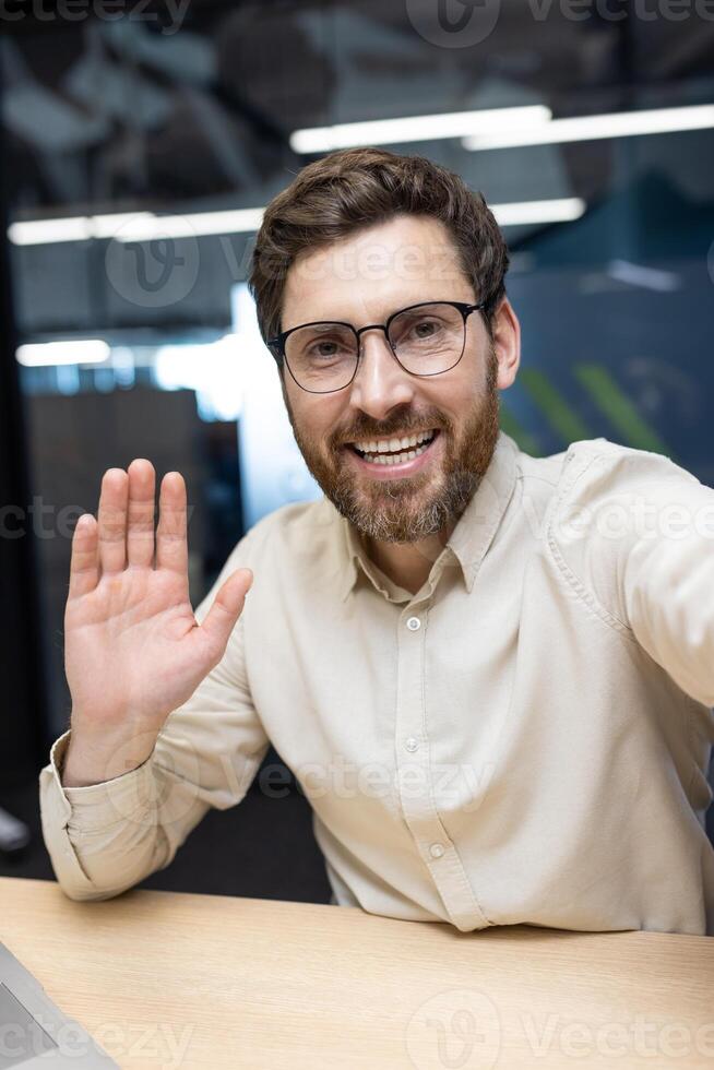 Vertical close-up photo of a young businessman in the office, talking on a call, greeting and waving at the camera.