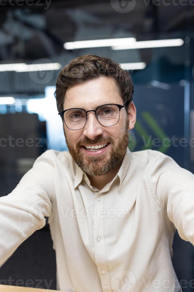 Vertical close-up photo of a young man in glasses and a shirt who is in the office and prays to the online camera he is holding in his hands.