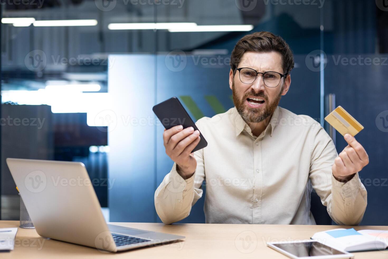 Portrait of an angry young man sitting at a desk in the office, holding a credit card and a mobile phone, looking at the camera in frustration, spreading his hands. photo
