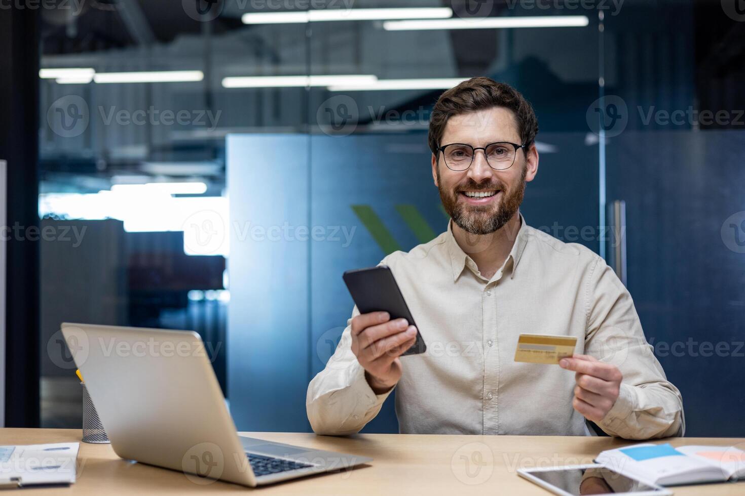 Close-up portrait of a young smiling businessman sitting at a desk in the office, holding a phone and a credit card in his hands, looking at the camera. photo