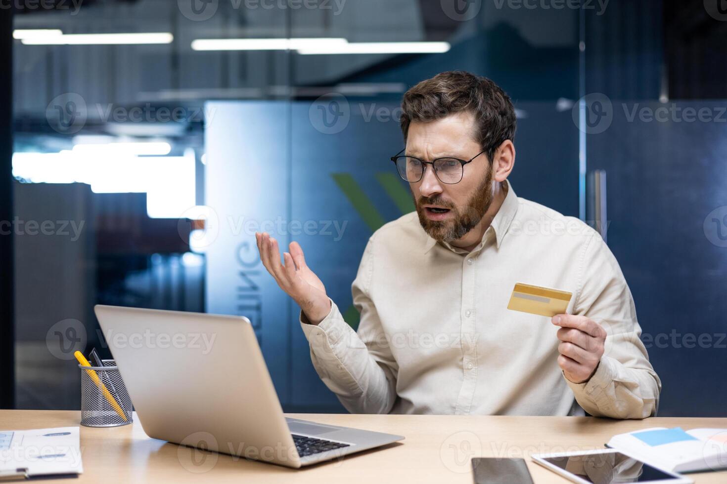 Financial problems. A young sad man is sitting at the desk in the office, holding a credit card in his hands and looking disappointedly at the laptop screen. photo