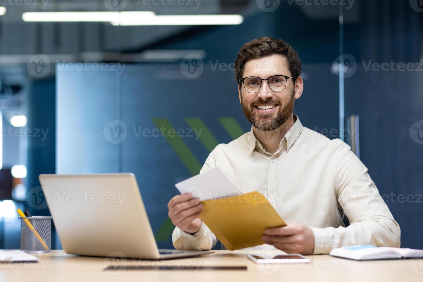 Portrait of a happy young businessman man who received good news and notification by letter, sitting in the office at the table, holding an envelope and looking at the camera. photo