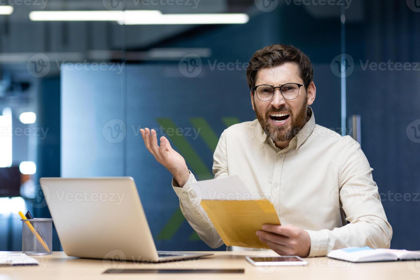 Angry young male businessman sitting in the office at the desk, holding an envelope with a received letter and looking disappointed at the camera. photo