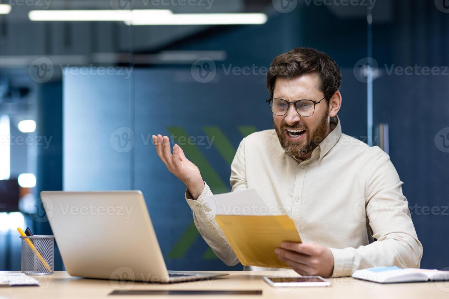 Angry young man businessman received a letter with bad news, sitting in the office at the table, holding an envelope in his hands and shouting unhappily. photo