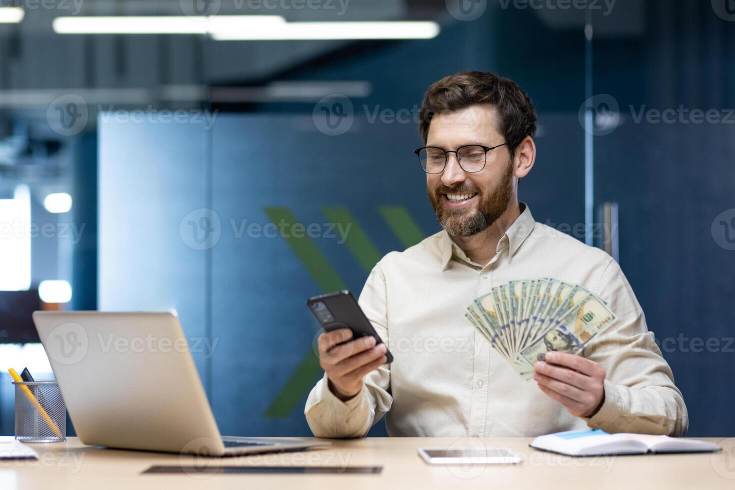 A happy young man is sitting in the office at the desk, holding a fan of cash money in his hands and using a mobile phone with a smile. photo