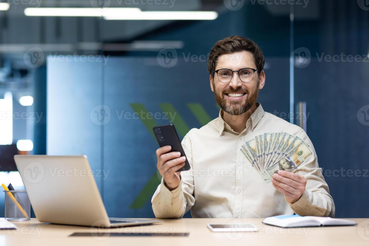 Portrait of a young businessman sitting in the office at a desk, holding a phone and a fan of dollar bills in his hands, smiling and looking at the camera. photo