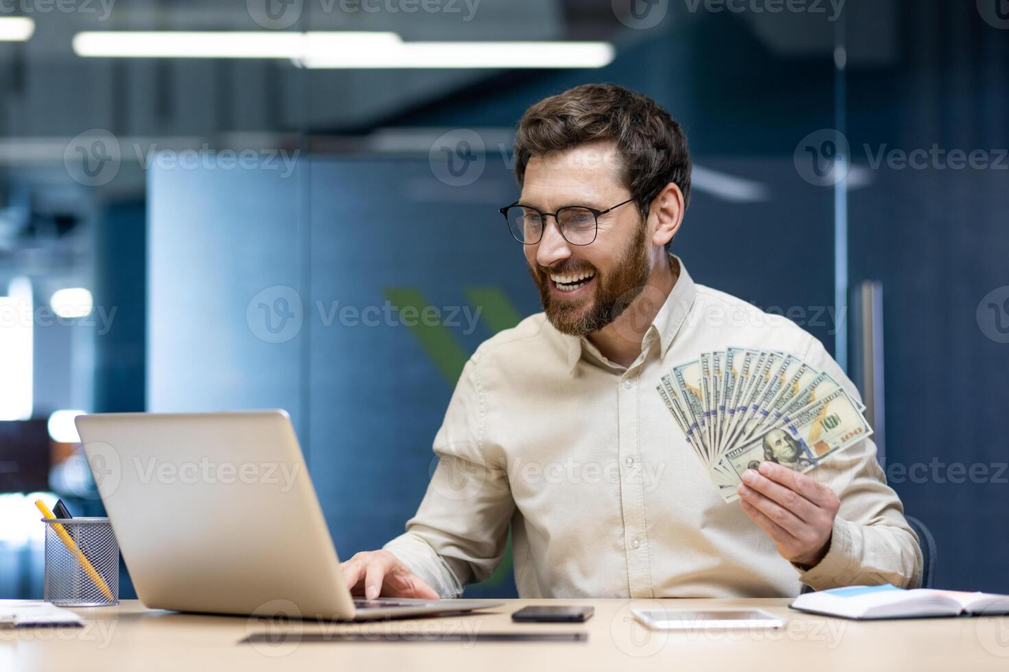 Satisfied and smiling young man sitting in the office at the desk, working on a laptop and holding a fan of money banknotes in his hand. photo