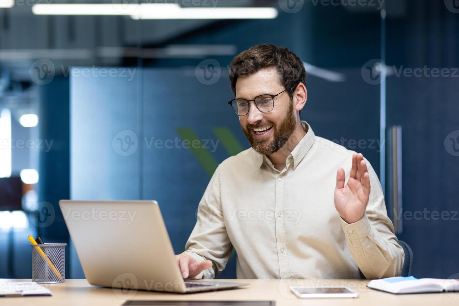 Smiling young man businessman sitting in office behind desk and talking on call on laptop, waving and greeting to camera. photo