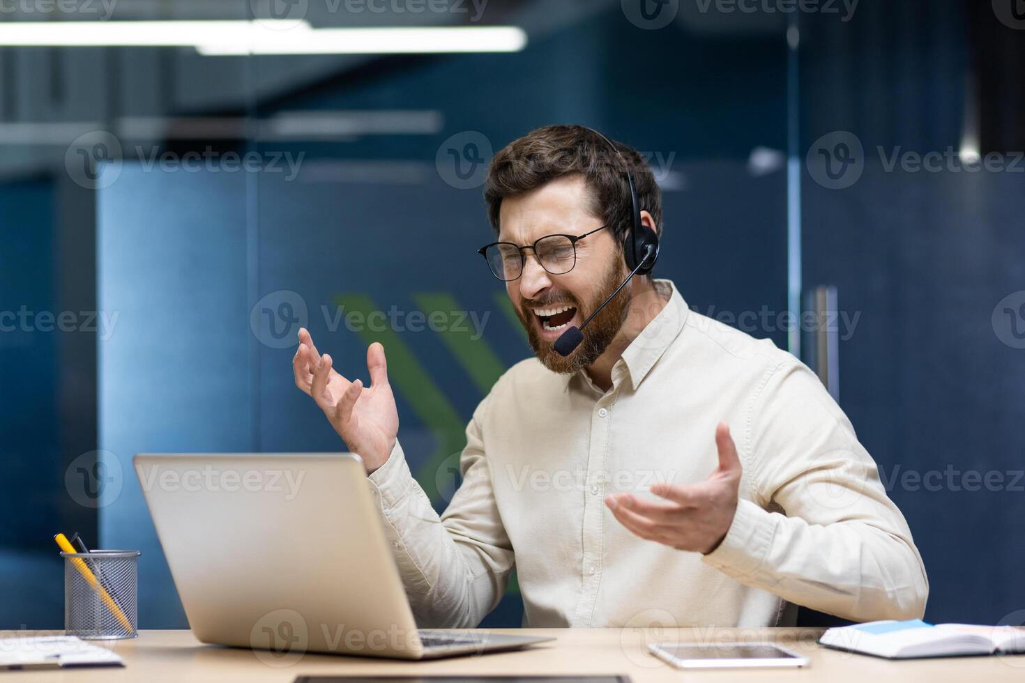 Young angry man in headset working in office on notebook, sitting at desk and emotionally shouting and arguing. photo