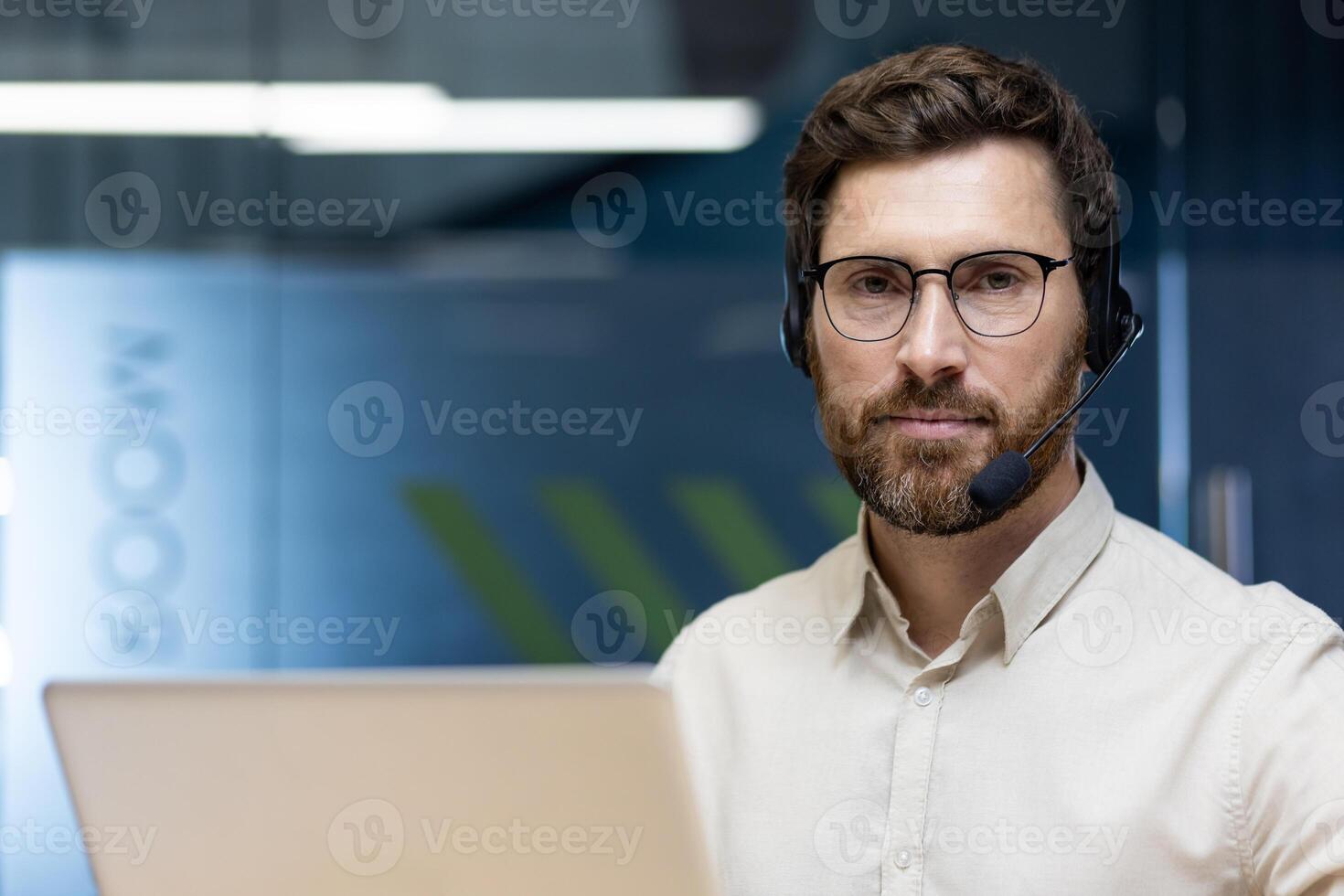 Close-up portrait of a young man in glasses and a headset sitting in the office at the workplace, working on a laptop, looking seriously into the camera. photo