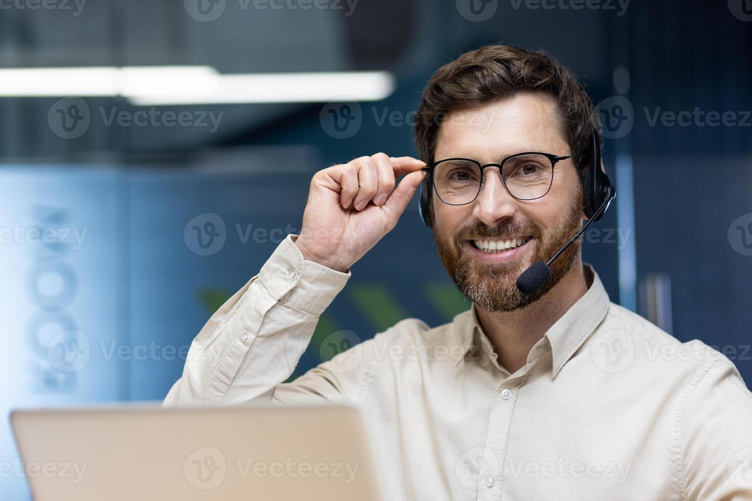 Close-up photo of a young man in a headset sitting at a desk in the front office, working on a laptop, smiling and looking at the camera while holding his glasses.