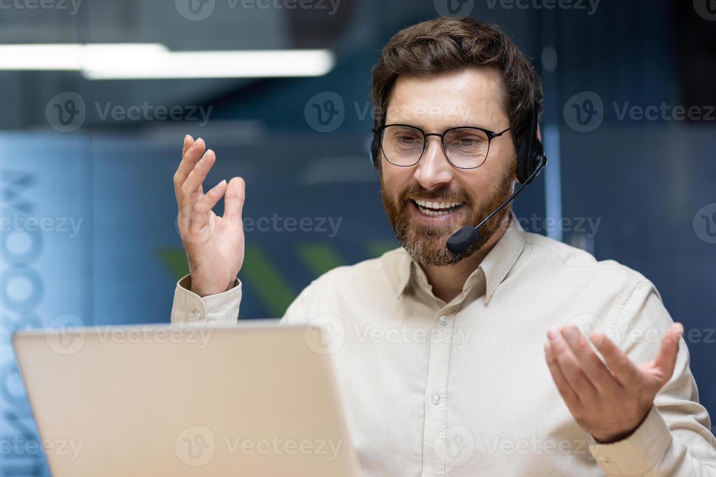 Close-up photo of a young smiling man in a headset sitting in the office at a table in front of a laptop and talking on a call.