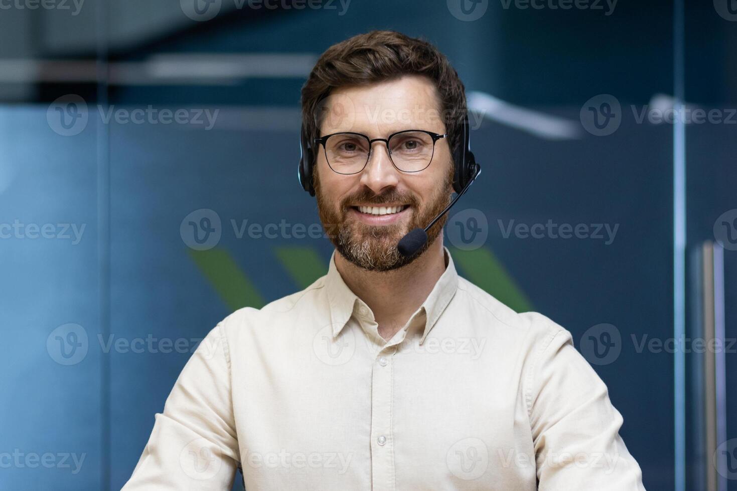 Close-up portrait of a young man in glasses wearing a headset sitting at a desk in an office and smiling at the camera. photo