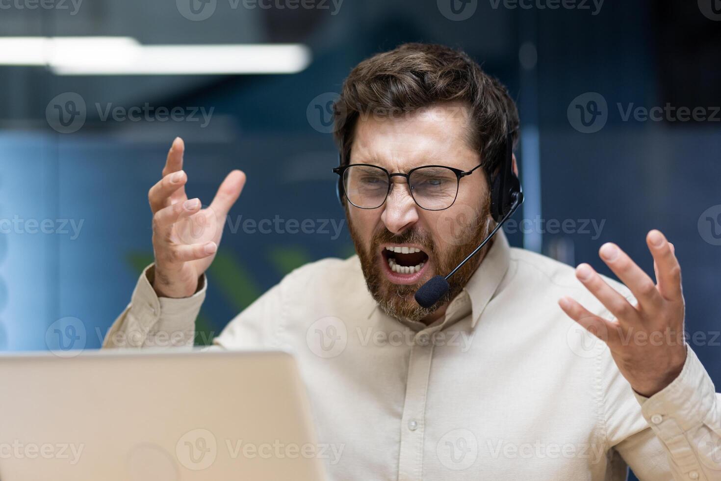 Close-up photo of young angry businessman man in headset shouting and arguing over call on laptop, aggressively gesturing with hands.
