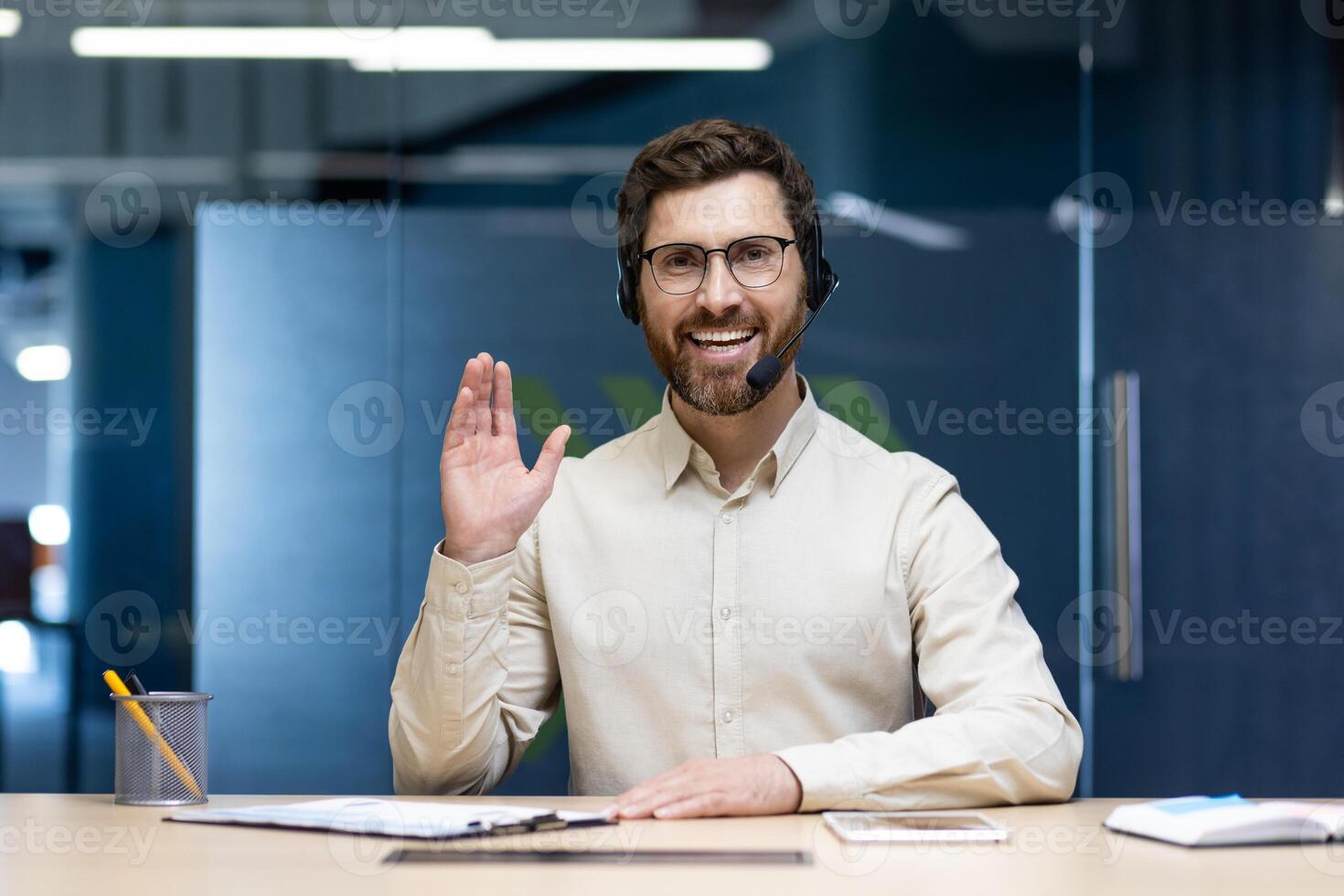 Portrait of a happy and successful young man in a headset sitting at a table in the office in front of the camera, smiling online, greeting and waving. photo
