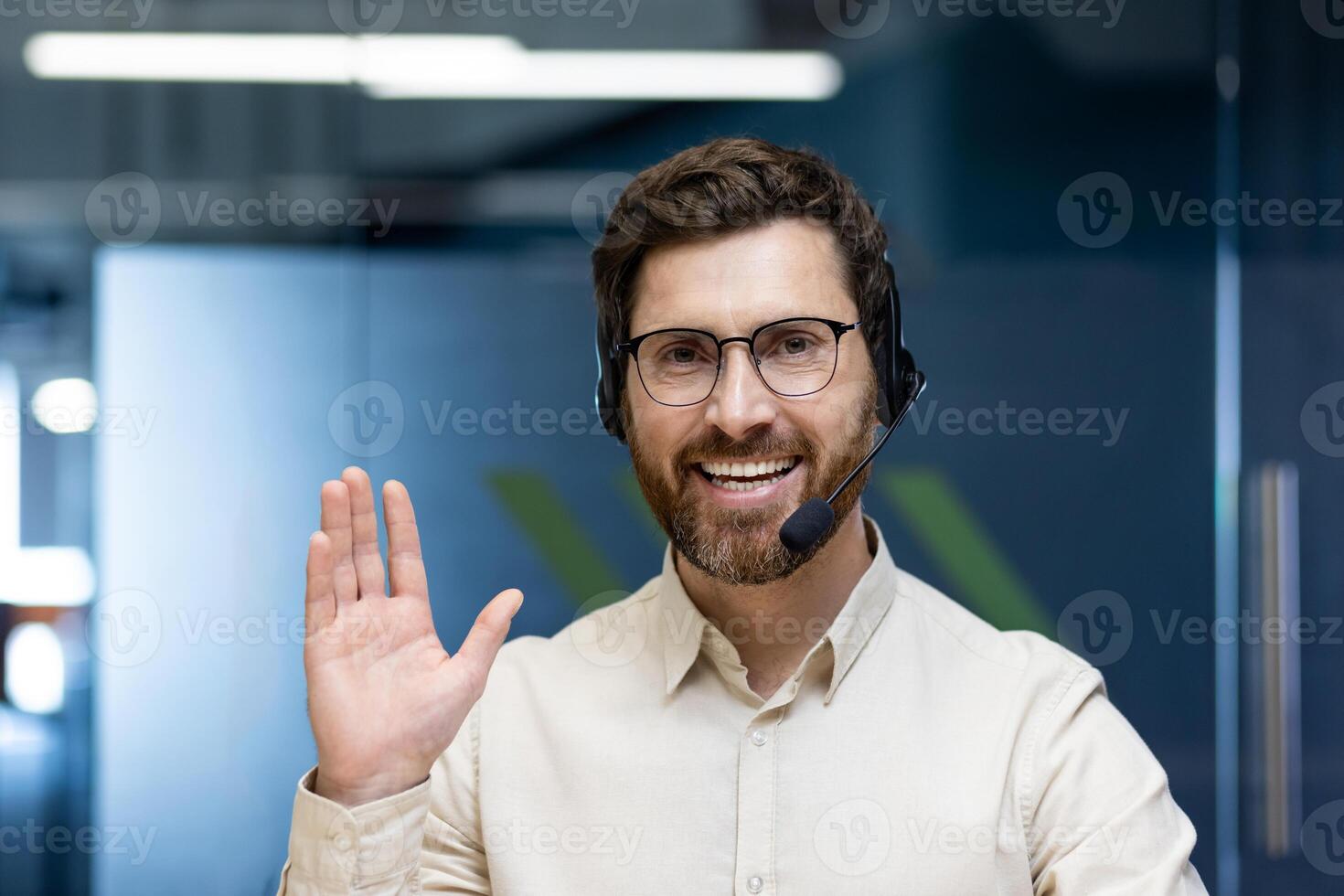Close-up portrait of a young man in a headset and glasses sitting in the office in front of the camera, greeting and waving. photo