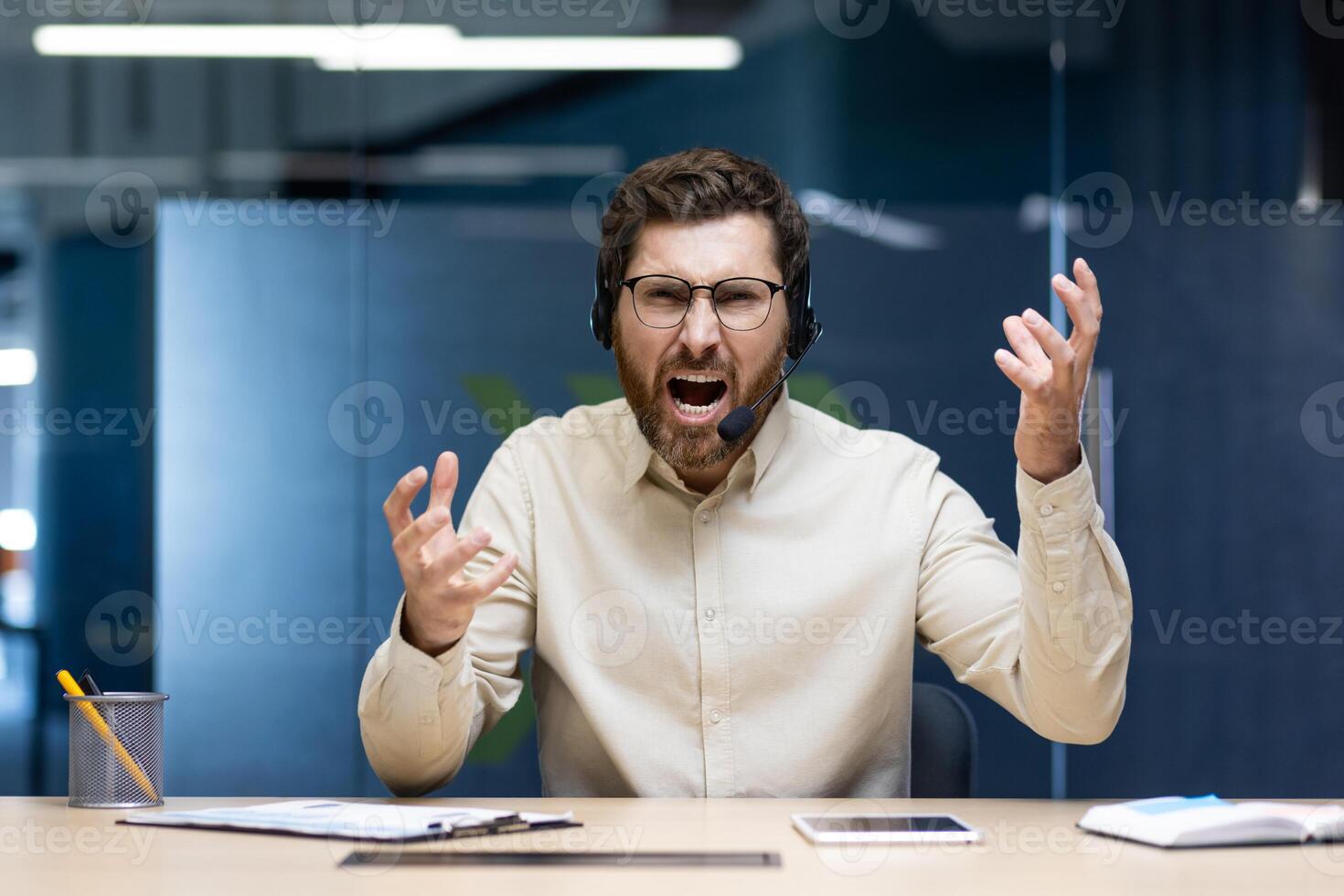Angry young man in a headset sitting in the office at the desk and aggressively shouting and talking to the camera while waving his hands. photo
