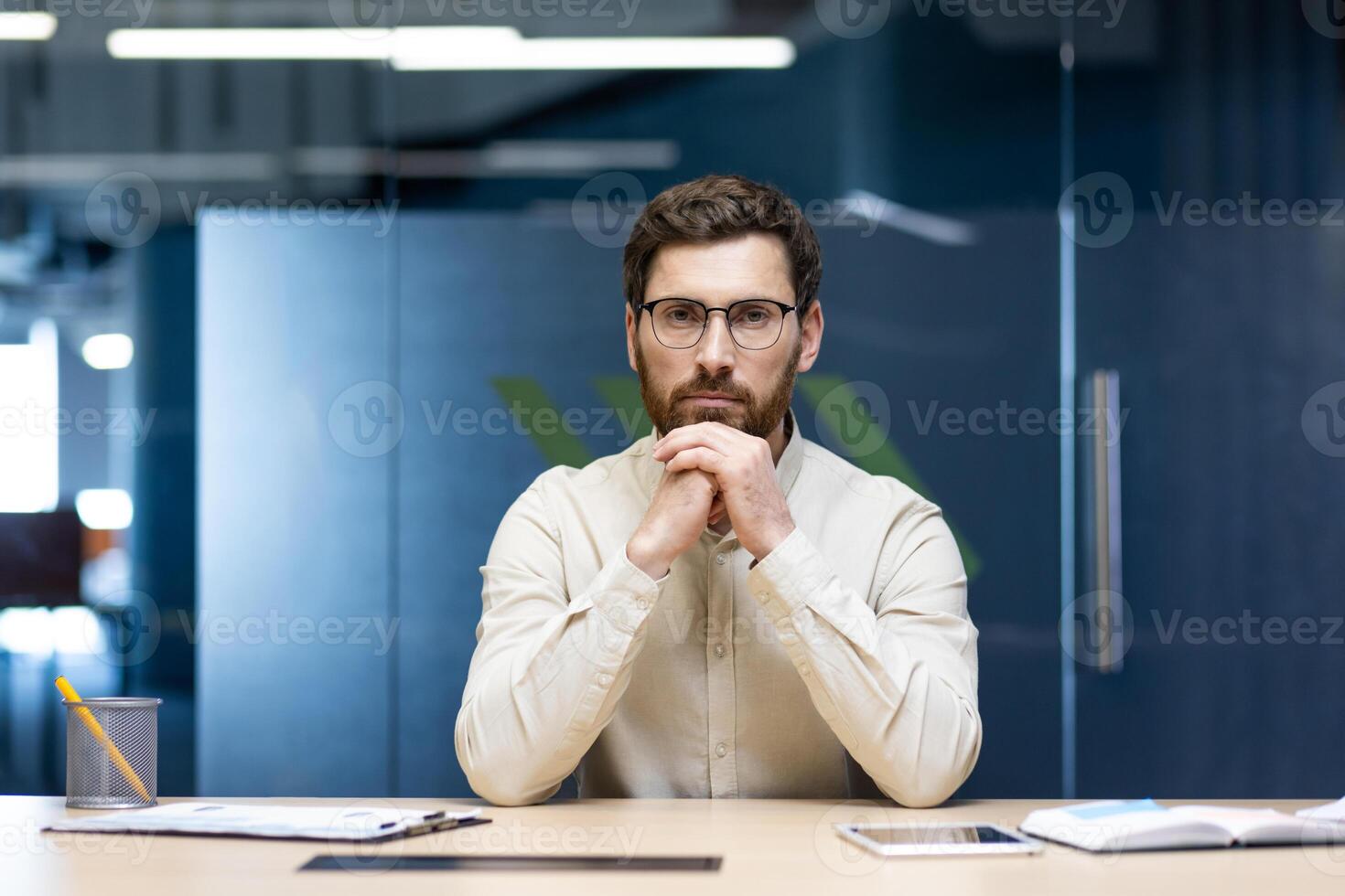 Close-up portrait of a young handsome businessman sitting at a desk in the office wearing glasses and a shirt, folded his hands together and confidently looking at the camera. photo