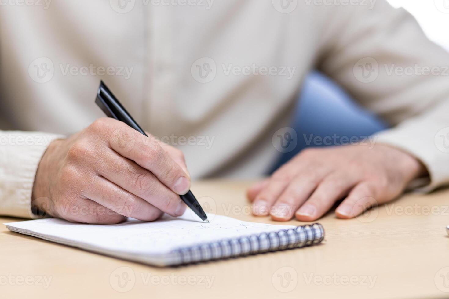 Close-up photo of a part of the body, hands of a young man in a light shirt holding a pen and making notes in a notebook at a desk in the office.