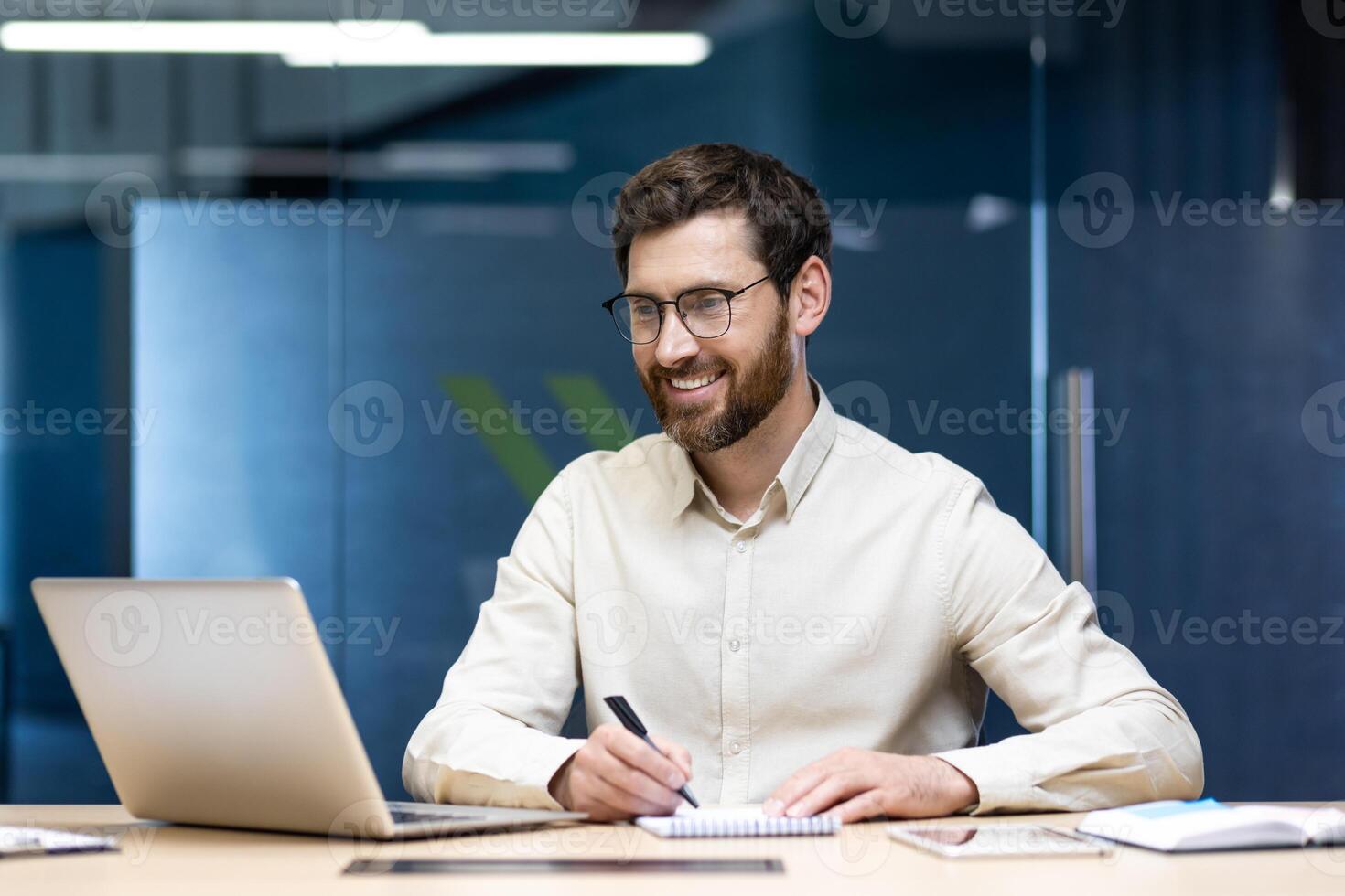 A smiling young man sits in the office at a desk in front of a notebook, makes notes in a notebook and looks at the screen with a smile. photo