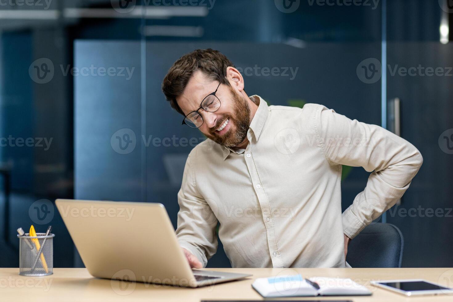 Tired young male office worker sitting at desk, holding hand behind back and suffering from lower back pain. photo