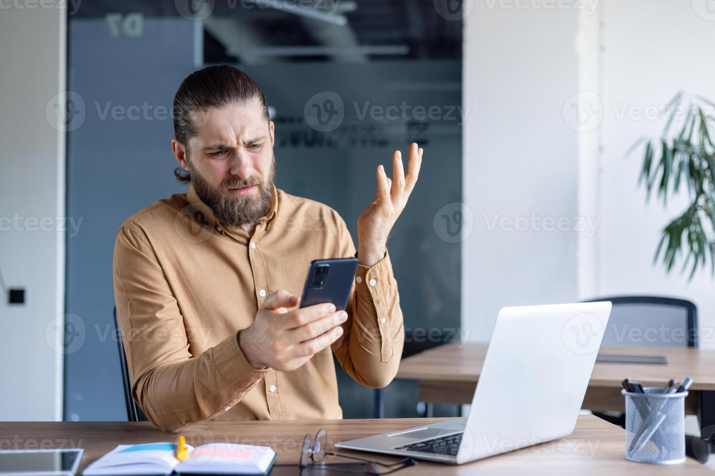 A frustrated man holds a smartphone, seemingly confused or exasperated while sitting at a desk. A laptop and office supplies are visible. photo
