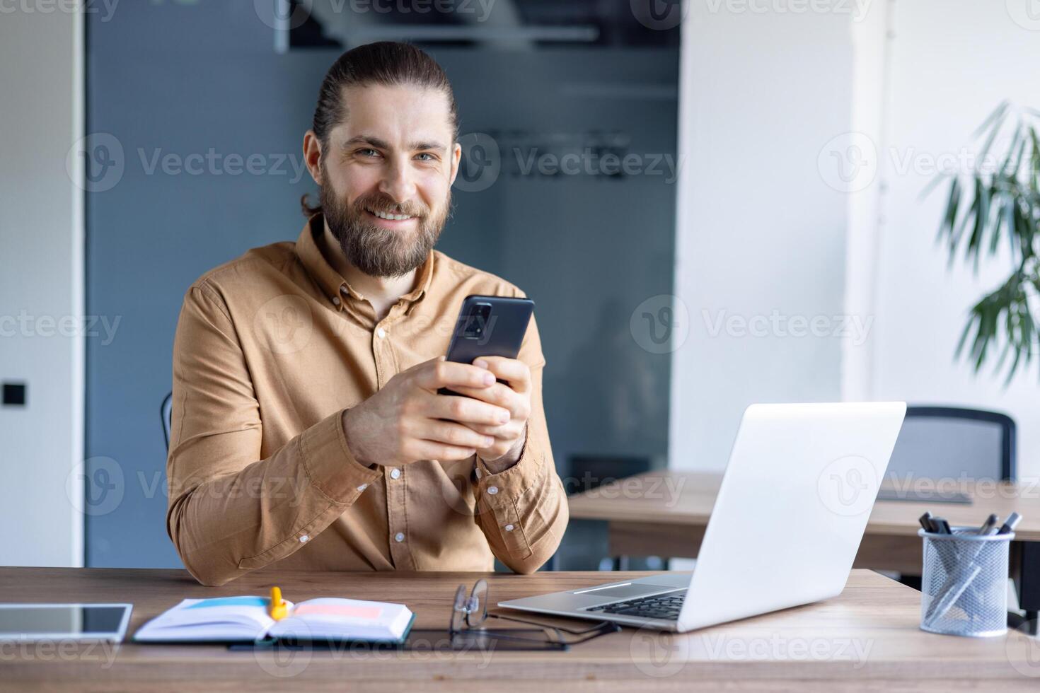 A smiling man with a beard uses his smartphone while sitting at his desk, focused and engaged. The open laptop hints at work. photo