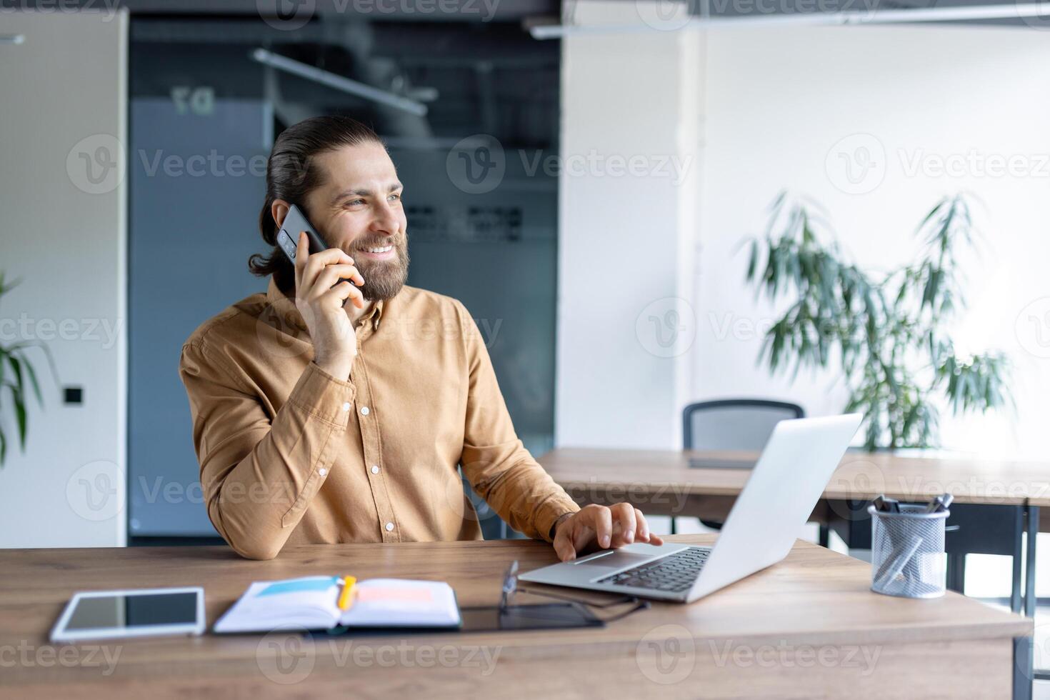 A man is smiling while talking on a smartphone in an office. He is working on a laptop at his desk in a modern office setting. photo