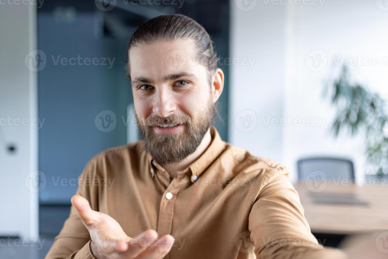 A man wearing shirt is smiling and gesturing with his hand during a call in a modern office setting. photo