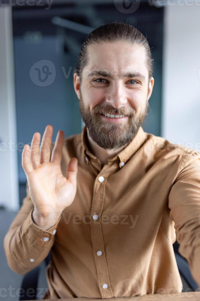 A bearded man waves at the camera, smiling. He wears a brown shirt and seems to be in a professional setting, possibly an office. photo