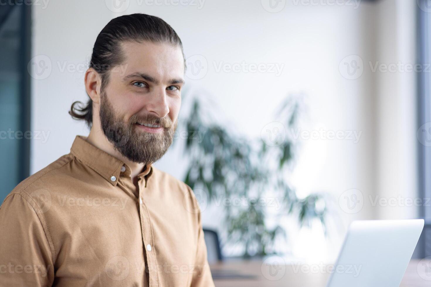 A smiling businessman with a beard looks towards the camera in an office setting. He is wearing a brown shirt and sitting near a laptop on a desk. photo
