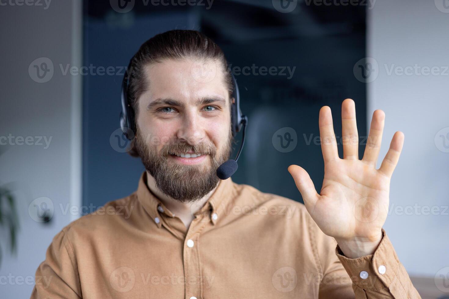 A smiling man with a headset waves at the camera, friendly and inviting. He is wearing a button-down shirt. photo